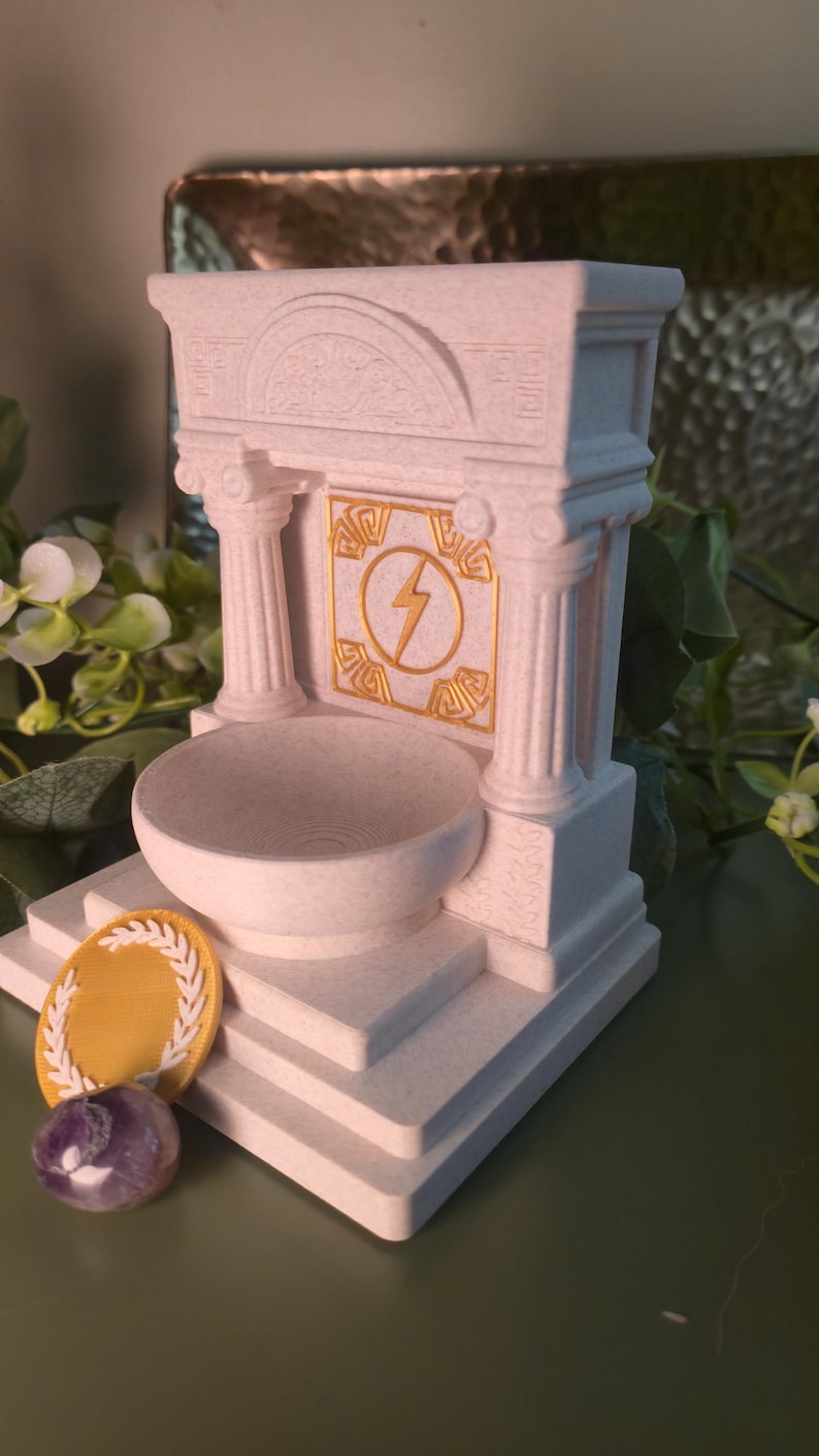 May include: A white, architectural-style decorative piece with a bowl, columns, and a lightning bolt emblem. A gold-colored laurel wreath and a purple stone are in front of the structure. The piece has a classical design.