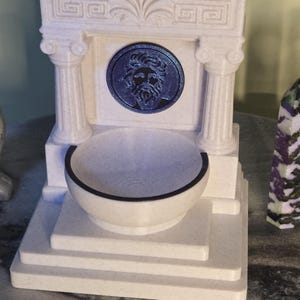 May include: A white, speckled, 3D-printed fountain featuring a classical architectural design with two columns, a pediment, and a central circular medallion depicting a bearded face in dark blue. A small, round basin sits at the base of the structure.