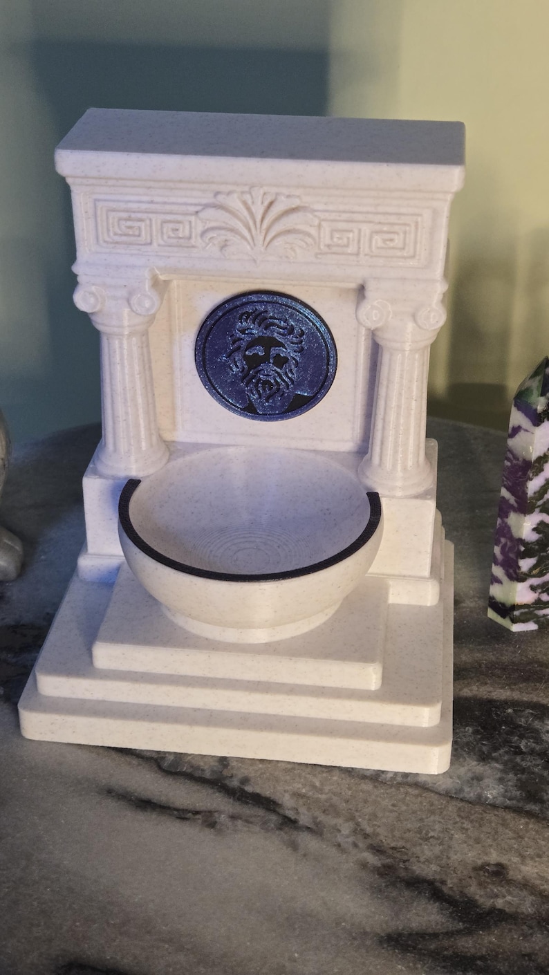 May include: A white, speckled, 3D-printed fountain featuring a classical architectural design with two columns, a pediment, and a central circular medallion depicting a bearded face in dark blue. A small, round basin sits at the base of the structure.