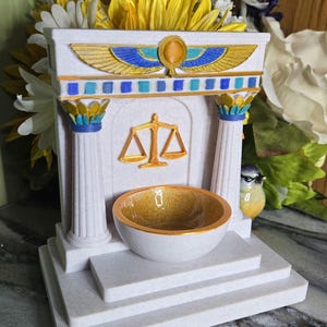 Può includere: Struttura decorativa bianca con accenti oro, blu e giallo. Presenta una ciotola, colonne e un simbolo della bilancia. La struttura è decorata con una composizione floreale sullo sfondo.