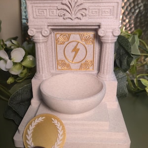 May include: A light-colored, miniature Greek-style temple with a round bowl and a gold lightning bolt emblem. A gold coin with a laurel wreath and a purple stone are in front of the temple. The temple has two columns.
