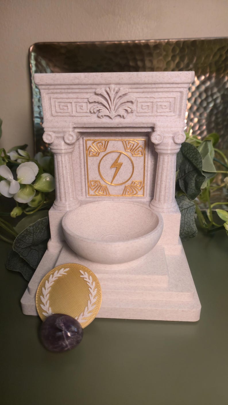 May include: A light-colored, miniature Greek-style temple with a round bowl and a gold lightning bolt emblem. A gold coin with a laurel wreath and a purple stone are in front of the temple. The temple has two columns.