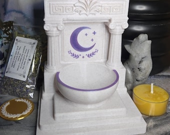 May include: A white, decorative incense burner with a purple crescent moon and star design. The burner has a bowl with a purple rim and is flanked by two columns. A yellow candle and a small bag of lavender are nearby.