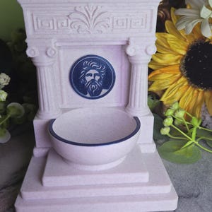 May include: A light pink speckled fountain-shaped object featuring two columns, a pediment with a carved fan design, and a central circular medallion depicting a bearded male face. A small basin with a dark rim sits at the base of the structure.
