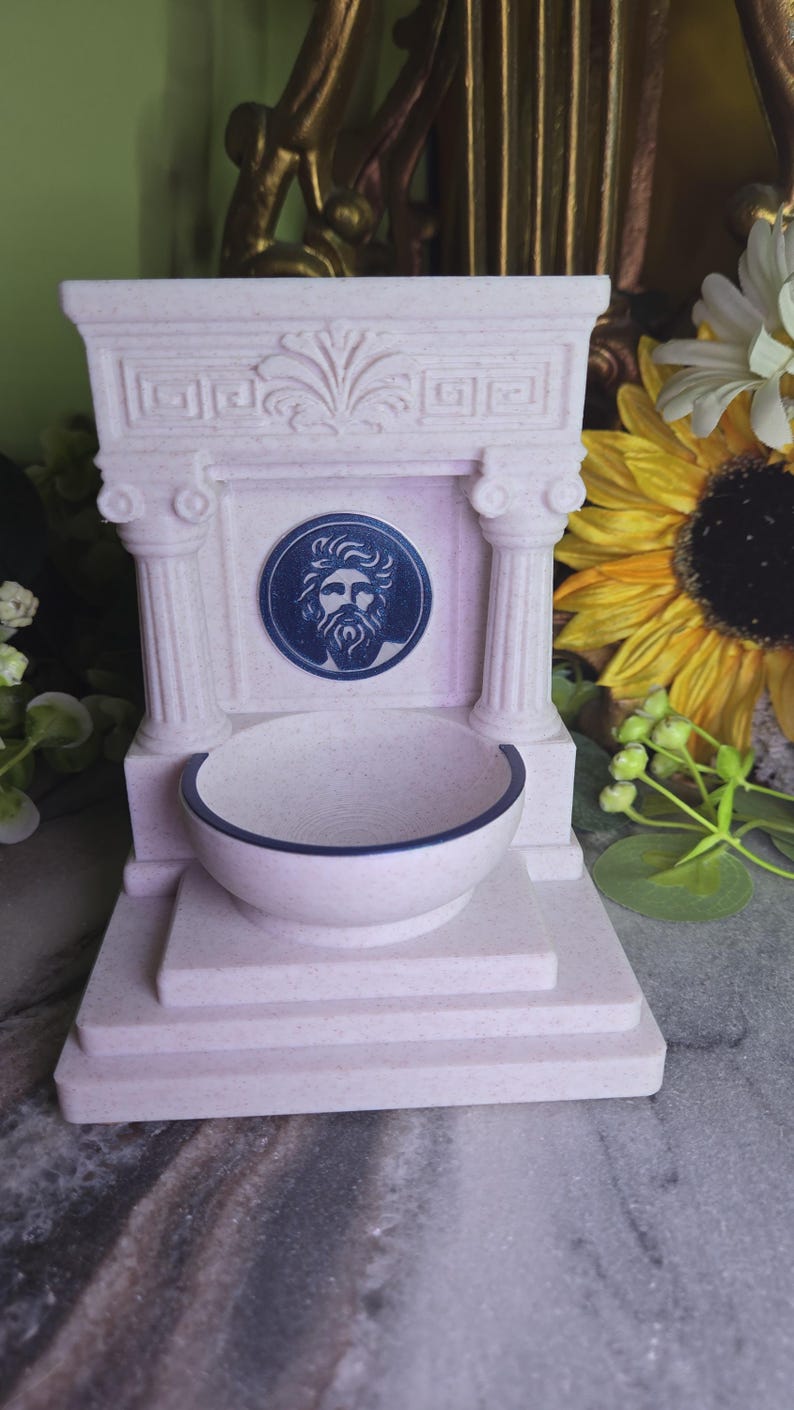 May include: A light pink speckled fountain-shaped object featuring two columns, a pediment with a carved fan design, and a central circular medallion depicting a bearded male face. A small basin with a dark rim sits at the base of the structure.