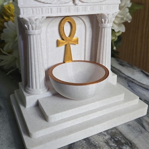 Puede incluir: Un altar decorativo blanco con un símbolo Ankh dorado y un cuenco pequeño. El altar presenta columnas y una base escalonada, con un borde dorado en el cuenco. El diseño recuerda la arquitectura del antiguo Egipto.