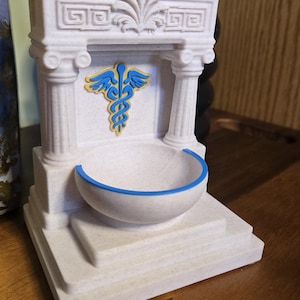 May include: A white decorative fountain with a blue and gold caduceus symbol. The fountain has a basin with a blue trim and is designed with classical architectural elements, including columns and decorative carvings.