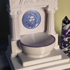 May include: A cream-colored, speckled fountain structure featuring two columns, a central circular medallion with a blue bearded face relief, and a basin with a purple rim. The architectural design includes a pediment with carved details.