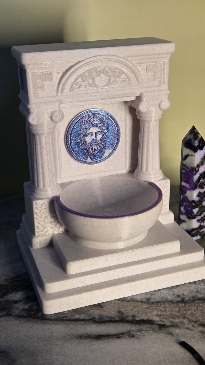 May include: A cream-colored, speckled fountain structure featuring two columns, a central circular medallion with a blue bearded face relief, and a basin with a purple rim. The architectural design includes a pediment with carved details.
