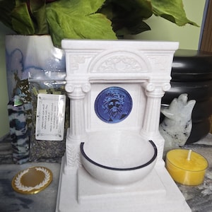 May include: A white, architectural-style fountain sculpture featuring two columns, a central blue circular medallion with a face, and a basin. The structure includes a small rectangular label with text, a gold coin, a yellow tea light, and a crystal.