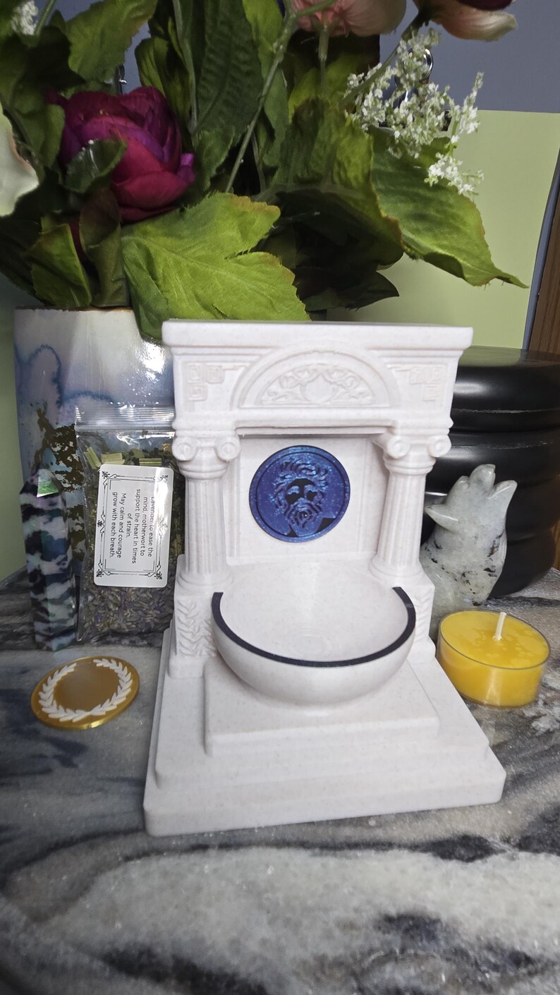 May include: A white, architectural-style fountain sculpture featuring two columns, a central blue circular medallion with a face, and a basin. The structure includes a small rectangular label with text, a gold coin, a yellow tea light, and a crystal.