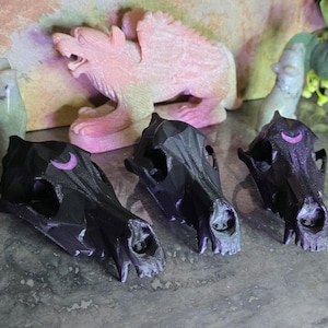 May include: Three black, low-poly animal skull figurines arranged in a row. Each skull features a small, purple crescent moon symbol on the forehead. The skulls have a faceted, geometric surface texture and are positioned on a grey surface.