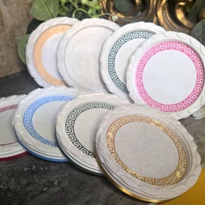 May include: A collection of round coasters featuring a white center and a raised, textured border. Each coaster displays a distinct colored Greek key pattern along the inner rim, including shades of pink, blue, green, red, gold, and white.