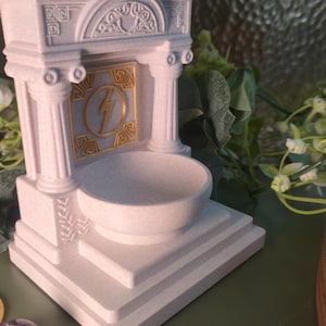 May include: A white, architectural-style incense holder with a bowl-shaped receptacle. The design includes columns, decorative carvings, and a gold-colored lightning bolt emblem. The holder is approximately 5 inches tall.