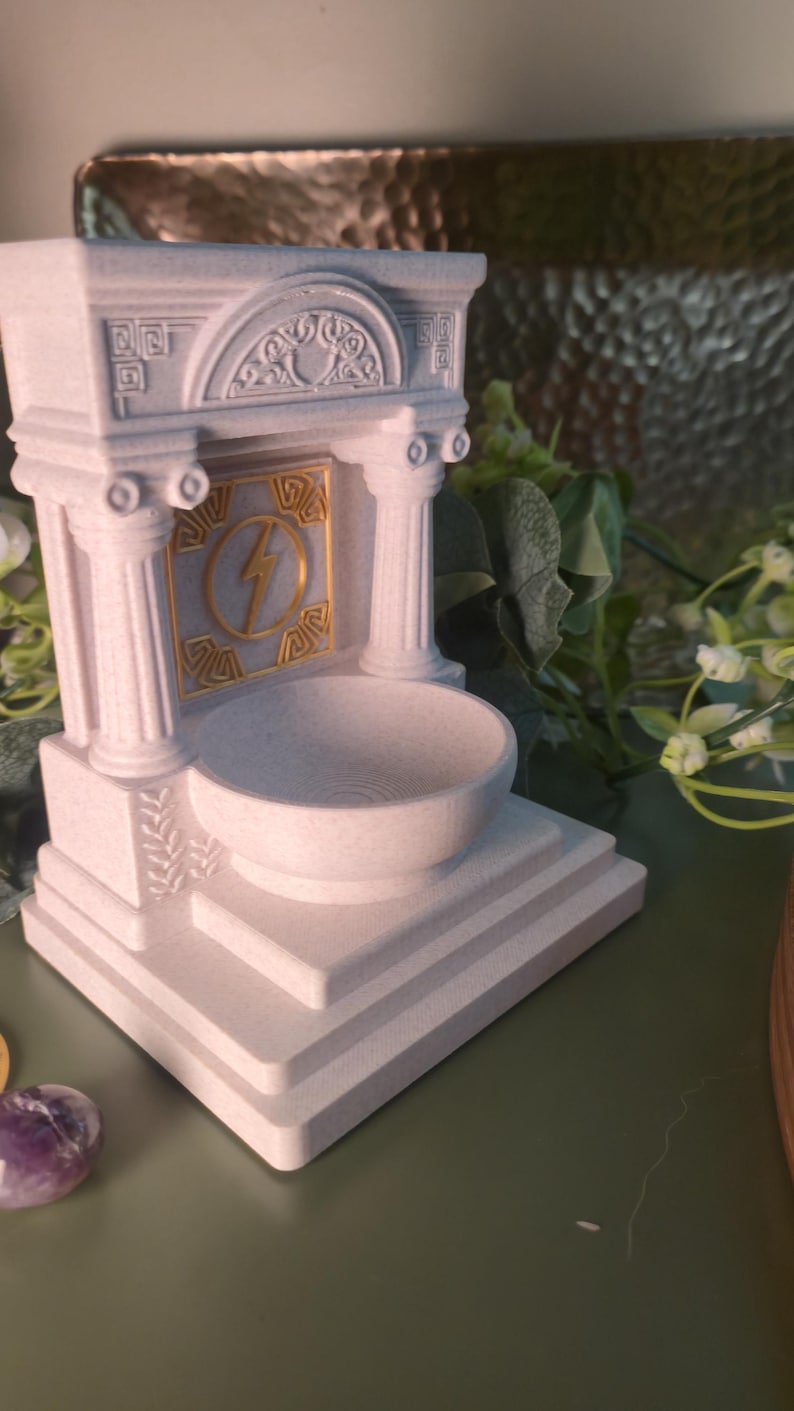 May include: A white, architectural-style incense holder with a bowl-shaped receptacle. The design includes columns, decorative carvings, and a gold-colored lightning bolt emblem. The holder is approximately 5 inches tall.