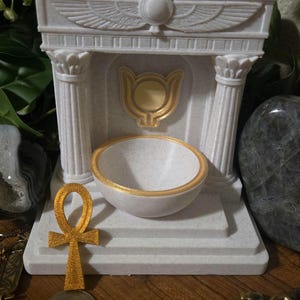 Puede incluir: Un altar decorativo blanco con detalles dorados. Presenta un cuenco con borde dorado, un ankh dorado y un emblema dorado. El diseño incluye columnas y detalles tallados, evocando el antiguo Egipto.