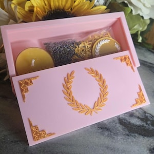 Könnte beinhalten: Eine rosa Box mit einem goldenen Lorbeerkranz-Design auf dem Deckel. Im Inneren sind eine gelbe Kerze, getrocknete Kräuter und eine goldfarbene Scheibe zu sehen. Die Box hat goldene Eckakzente und ist geöffnet, um den Inhalt zu zeigen.