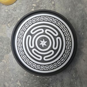 May include: A circular black tray featuring a white printed design. The center displays a stylized labyrinthine pattern surrounding a six-pointed star. The outer border is decorated with a continuous white Greek key meander pattern.