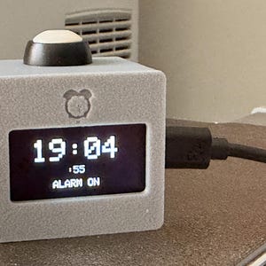 Op de afbeelding: Een grijze kubusvormige digitale wekker met een zwart scherm dat de tijd 19:04, seconden en de tekst ALARM ON weergeeft. Bovenop zit een zwarte knop en aan de rechterkant van het apparaat is een zwarte stroomkabel aangesloten.