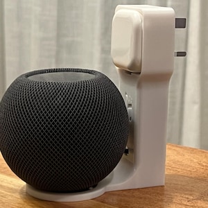 Puede incluir: Un altavoz inteligente negro con un diseño de malla se encuentra sobre una base blanca. Un adaptador de corriente blanco con un enchufe está colocado junto al altavoz. El altavoz y el adaptador están sobre una superficie de madera.