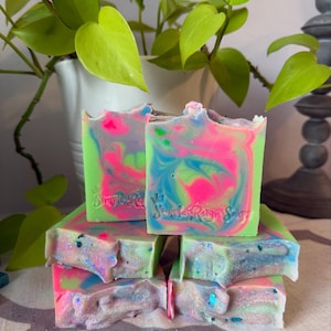 May include: A stack of handmade soap bars with vibrant colors. The soap features swirls of neon pink, blue, and green, with a textured surface and glitter accents. The soap is branded with the text "Sweet Reign Soap".