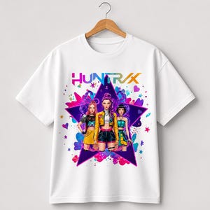 May include: White t-shirt with a graphic of three stylized figures in front of a purple star. The word "HUNT/RX" is printed above the figures in a colourful font. The design includes hearts and other decorative elements.