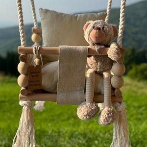 Op de afbeelding: Een houten babyschommel met een beige teddybeer en een beige kussen. De schommel hangt aan een dik, wit touw en heeft decoratieve houten kralen. De schommel heeft een beige stoffen zitting en een label met tekst.