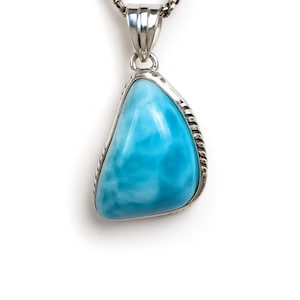 Puede incluir: Un collar de plata con un colgante en forma de pera. El colgante presenta una piedra preciosa azul claro con un efecto veteado. La piedra está enmarcada por un borde plateado y cuelga de una cadena plateada.