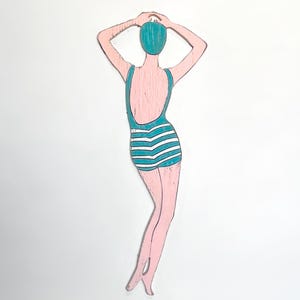 May include: Wooden cutout of a woman in a vintage swimsuit. The figure is painted pink with a teal swimsuit and swim cap. The swimsuit has white horizontal stripes. The woman is posed with her arms above her head.