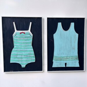 Pu&ograve; includere: Due opere d'arte in legno incorniciate. Una raffigura un costume da bagno a righe turchese e azzurre con un fiocco rosso. L'altra &egrave; un costume da bagno azzurro con pantaloncini. Entrambe sono su sfondo blu scuro.