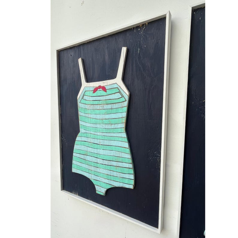 Pu&ograve; includere: Opera d'arte in legno di un costume da bagno in stile vintage. Il costume da bagno &egrave; azzurro con strisce orizzontali e un fiocco rosso. &Egrave; ambientato su uno sfondo nero all'interno di una cornice bianca. Un pezzo unico di decorazione murale.