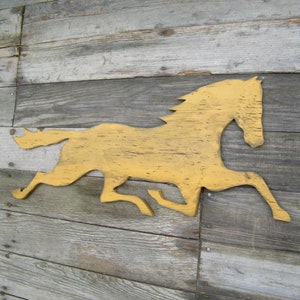May include: A wooden cutout of a horse running, painted in a bright yellow colour. The horse is mounted on a weathered wooden background.