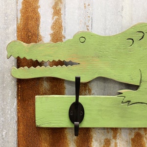 May include: Green wooden alligator head wall hook with a black metal hook. The alligator head is painted in a distressed green paint with a light brown undercoat. The hook is attached to a green wooden board.