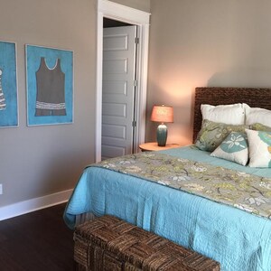 Pu&ograve; includere: Camera da letto con copriletto azzurro, cuscini decorativi e testiera intrecciata. Due opere d'arte incorniciate di costumi da bagno vintage sono appese alla parete. Una lampada &egrave; su un comodino e una panca intrecciata ai piedi del letto.