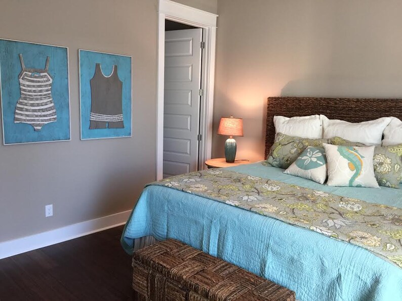 Pu&ograve; includere: Camera da letto con copriletto azzurro, cuscini decorativi e testiera intrecciata. Due opere d'arte incorniciate di costumi da bagno vintage sono appese alla parete. Una lampada &egrave; su un comodino e una panca intrecciata ai piedi del letto.