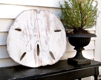 Può includere: Decorazione da parete in legno di dollaro di sabbia sbiancato con un motivo floreale intagliato. Il dollaro di sabbia è appeso a una parete bianca con una panca nera e una pianta in vaso sullo sfondo.