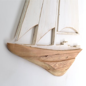 May include: Wooden sailboat wall art with a distressed white finish on the sails and a natural wood hull. The nautical-themed decor piece is designed to be mounted on a wall, adding a coastal touch to any room.