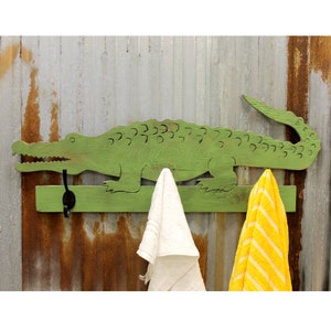 May include: Green wooden alligator shaped towel rack with two hooks. The rack is mounted on a wall and has two towels hanging from it, one white and one yellow with stripes.