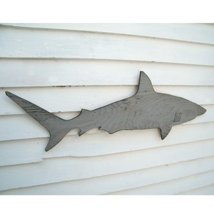 May include: A gray wooden shark silhouette mounted on a white wooden wall. The shark has a dorsal fin, a tail fin, and a detailed body shape. The wood grain is visible, adding texture to the artwork. This nautical decor piece is suitable for a beach house or coastal theme.