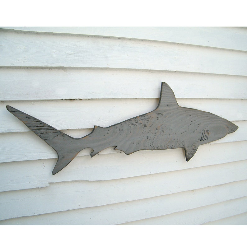 May include: A gray wooden shark silhouette mounted on a white wooden wall. The shark has a dorsal fin, a tail fin, and a detailed body shape. The wood grain is visible, adding texture to the artwork. This nautical decor piece is suitable for a beach house or coastal theme.