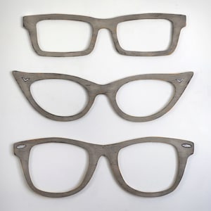 May include: Three wooden frames in the shape of eyeglasses. The frames are all different styles, with the top frame being a classic rectangular shape, the middle frame being a cat-eye shape, and the bottom frame being a more modern rounded shape.