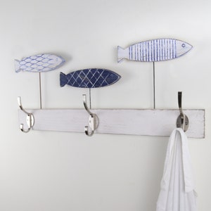 May include: White wooden coat rack with three hooks and three blue and white fish-shaped hooks. A white towel is hanging on one of the hooks.