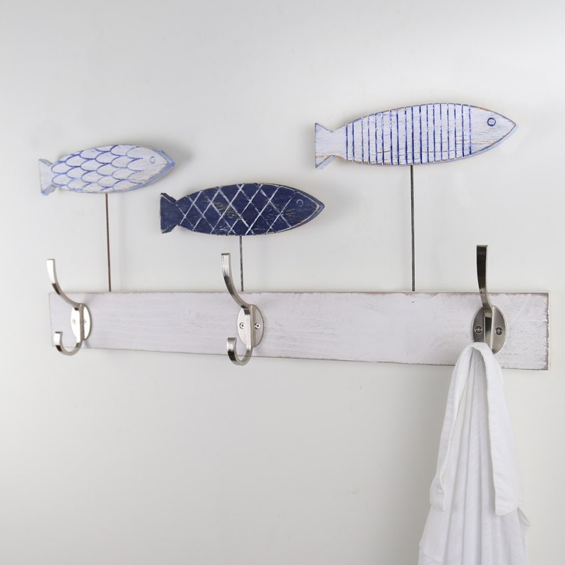 May include: White wooden coat rack with three hooks and three blue and white fish-shaped hooks. A white towel is hanging on one of the hooks.