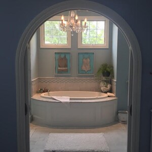 Pu&ograve; includere: Un bagno azzurro con una vasca da bagno bianca, un tappeto bianco e un lampadario di cristallo. Due opere d'arte incorniciate di costumi da bagno sono appese alla parete sopra la vasca. La stanza &egrave; vista attraverso una porta ad arco.