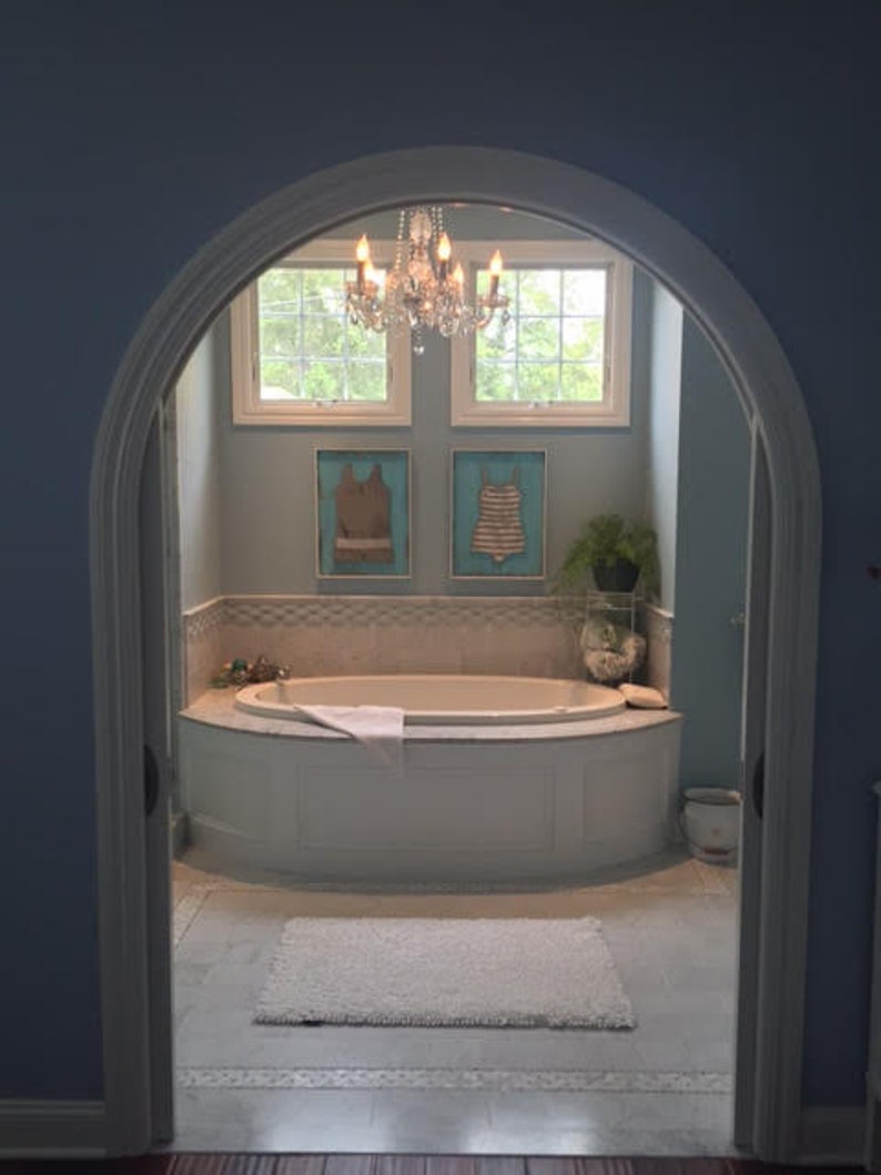 Pu&ograve; includere: Un bagno azzurro con una vasca da bagno bianca, un tappeto bianco e un lampadario di cristallo. Due opere d'arte incorniciate di costumi da bagno sono appese alla parete sopra la vasca. La stanza &egrave; vista attraverso una porta ad arco.