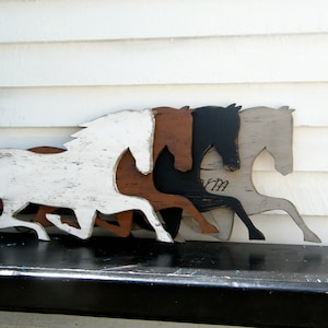 May include: Four wooden horse silhouettes in various shades of white, brown, black, and grey, arranged in a row on a black surface. The horses are facing the right side of the image and appear to be running.