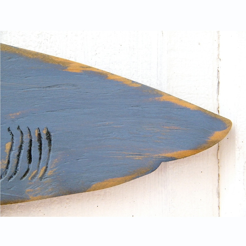 May include: A wooden shark fin sculpture painted in a distressed style. The fin is primarily blue-gray with a weathered, golden-brown edge. The shark's gills are carved into the wood. The background is a white, wooden surface.