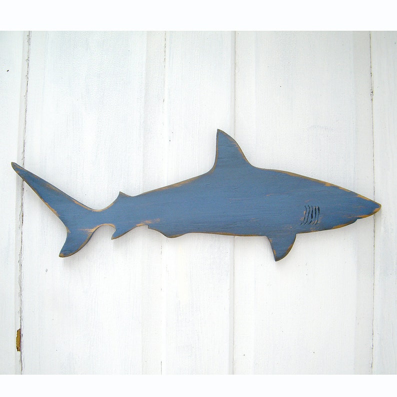 May include: A blue wooden shark cutout with a distressed finish. The shark has a dorsal fin, tail fin, and gills carved into the wood. The shark is mounted on a white wooden background.