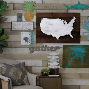 May include: A wall decor set with a nautical theme. The set includes a wooden sign with the word "gather" in metal letters, a wooden sign with a blue silhouette of a palm tree, a wooden sign with a blue silhouette of a crab, a wooden sign with a blue silhouette of a shark, a wooden sign with a white silhouette of the United States, a wooden sign with a blue silhouette of a seagull, and a wooden sign with a blue and white silhouette of a pineapple.