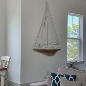 May include: A white sailboat wall decor with a wooden base is mounted on a white wall. The sailboat has a simple design with a mast and sail. The room also features a gray sofa and decorative pillows.